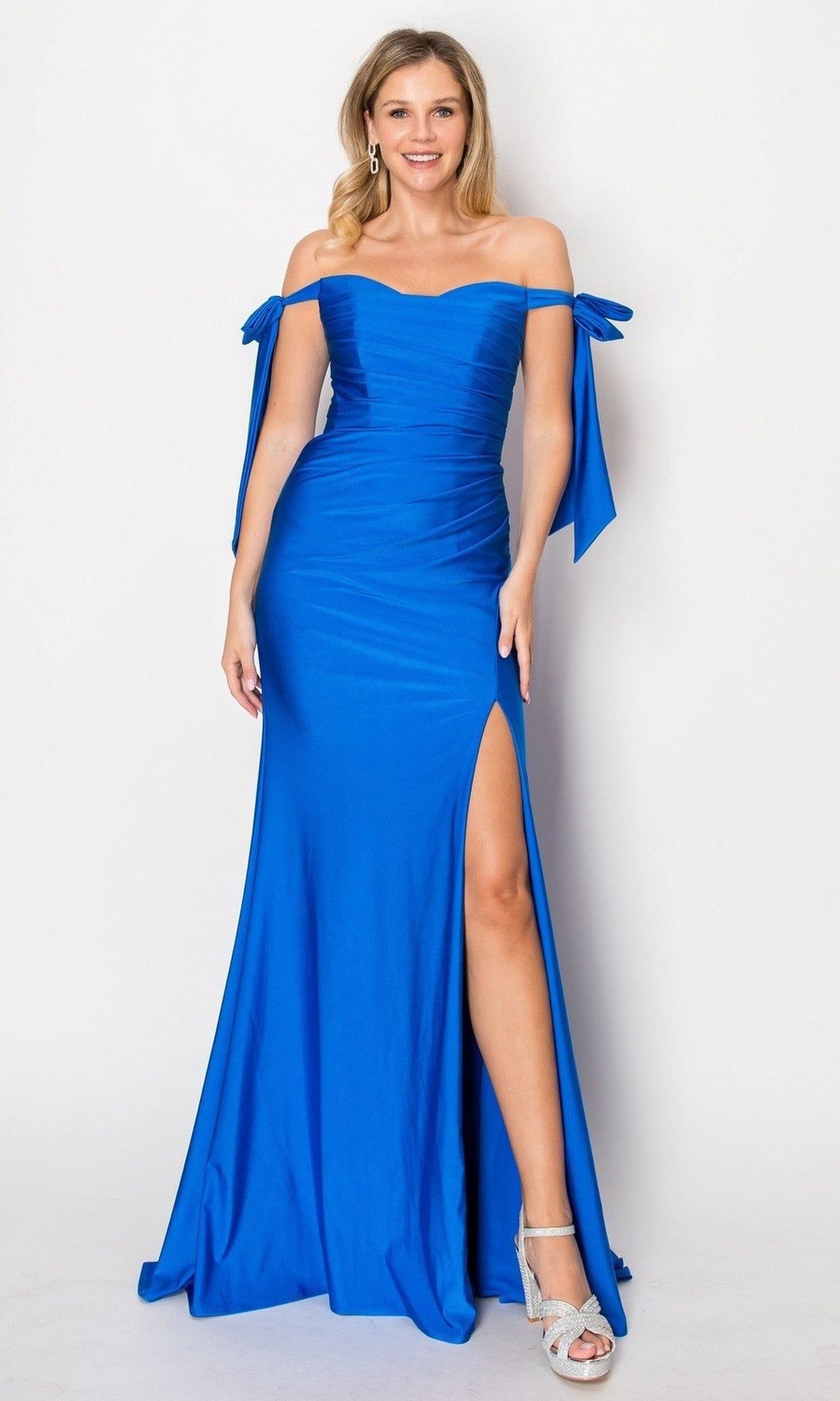 A woman wearing the Long Prom Dress: Chicas DM8010 in royal, an elegant off-the-shoulder gown featuring a thigh-high slit and bow details on the sleeves, paired with silver high-heeled sandals, stands against a plain white background.
